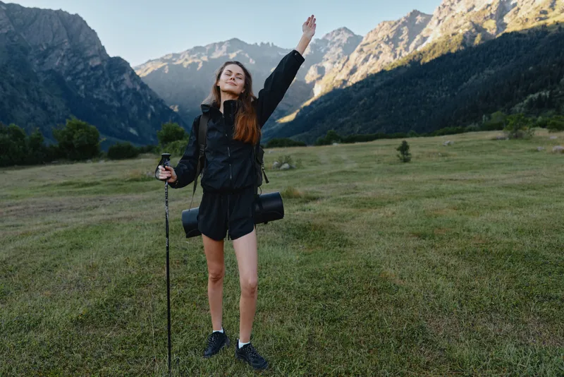 A joyful hiker woman stands in a mountain meadow with trekking poles, smiling at the camera. She appears energized and ready for an outdoor adventure, conveying a fresh, confident, and active