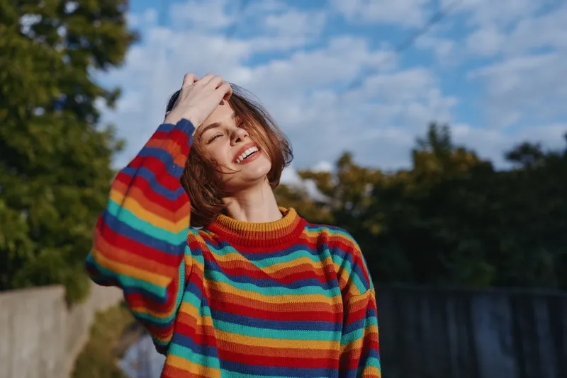 Young woman smiling in a rainbow sweater outdoors in a park, casual outfit, mid twenties model with hand on head, sunny natural light, relaxed lifestyle portrait for fashion and travel.