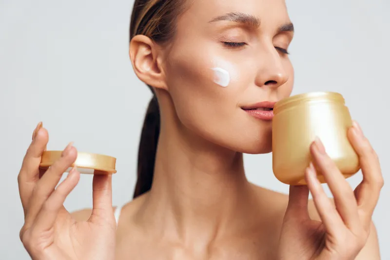 A young woman applies moisturizer from a gold jar, patting cream onto her cheek in a soft studio setting, illustrating skincare routine and self care.