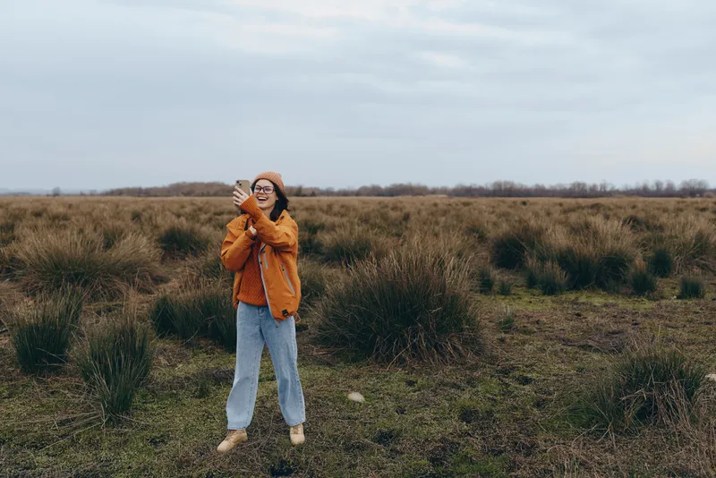 Woman lifestyle portrait outdoors in grassy field on cloudy day, orange jacket and blue jeans, smiling and holding smartphone, standing on dirt path with meadow background, natural light