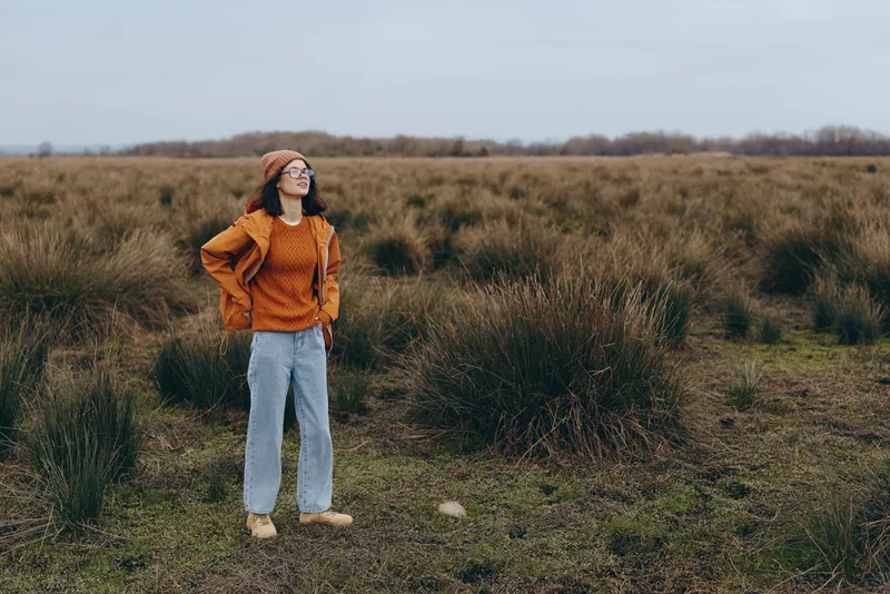 Woman lifestyle outdoors, standing in grassy field with natural daylight, wearing orange sweater, blue jeans, white shoes, sunglasses, hands in pockets, calm casual pose, confident