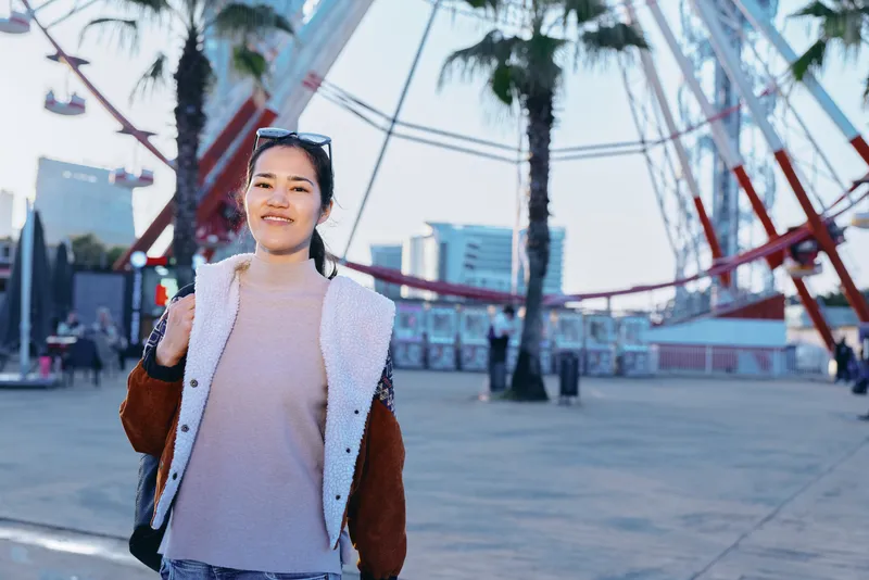 Smiling woman in a light sweater and varsity jacket walks along a sunny boardwalk with palm trees and a ferris wheel in the background, urban casual fashion and travel vibe.