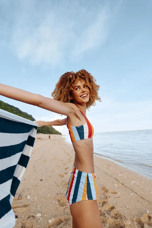 Young woman with curly hair enjoys a sunny day on the beach wearing a colorful striped swimsuit while holding a striped towel behind her back with a joyful expression.