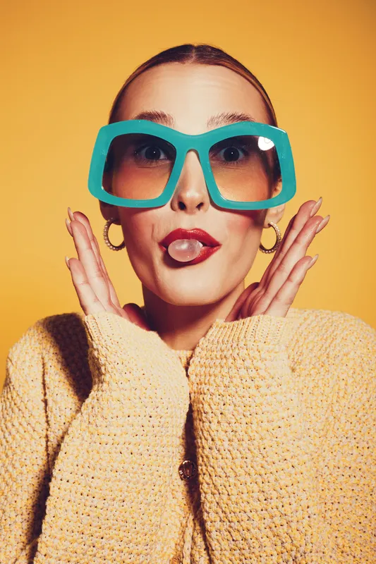 Young woman with oversized blue sunglasses makes funny face with tongue out and hands near face. Bright yellow background and cozy knitted sweater create playful mood.