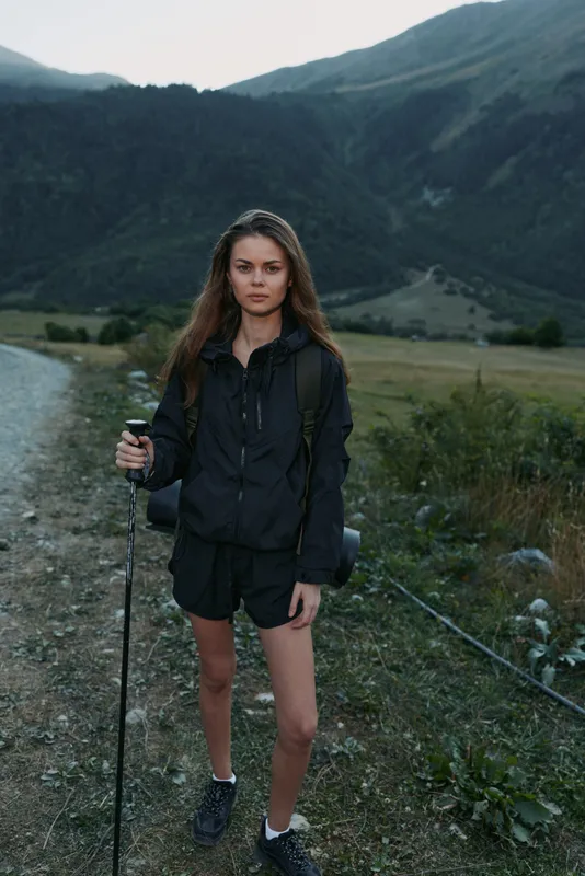 A young female hiker stands on a rugged trail in a mountainous landscape, holding a trekking pole. She wears a dark jacket, shorts, and a backpack, exuding readiness, focus, and curiosity.