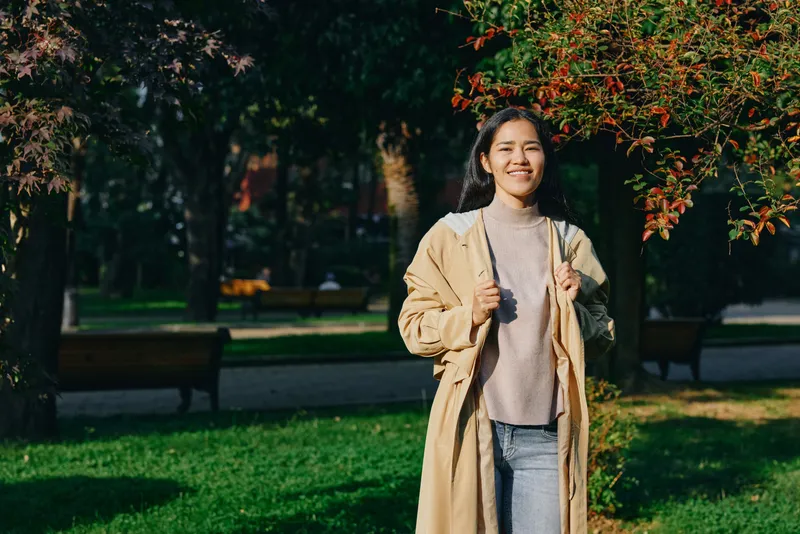 Woman in a beige coat strolling through a sunlit park, autumn trees glow with warm light, casual sweater and relaxed mood, outdoor fashion storytelling with greenery, benches and open path for