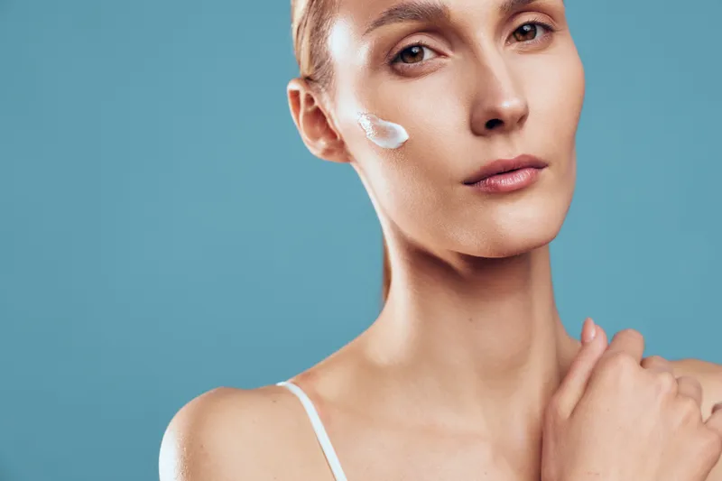 Young woman with fair skin applies moisturizer on her cheek while looking thoughtfully at the camera against a plain blue background in a skincare routine moment.
