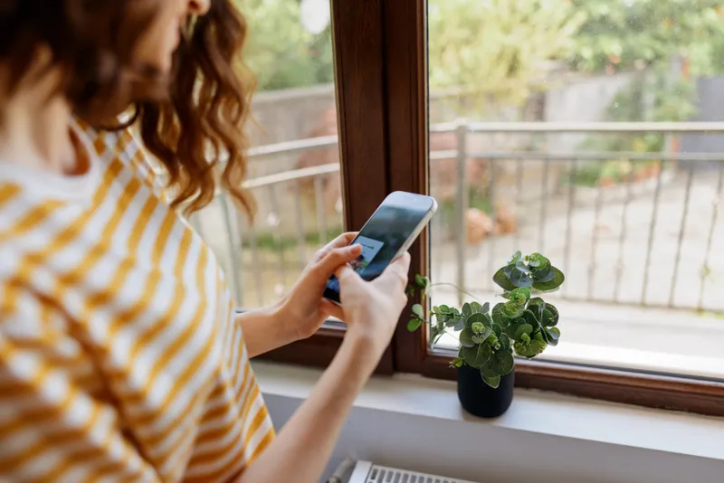 Woman in striped shirt using smartphone by a bright window, with a small plant on the sill. Casual home lifestyle scene highlighting daily tech use and self care.