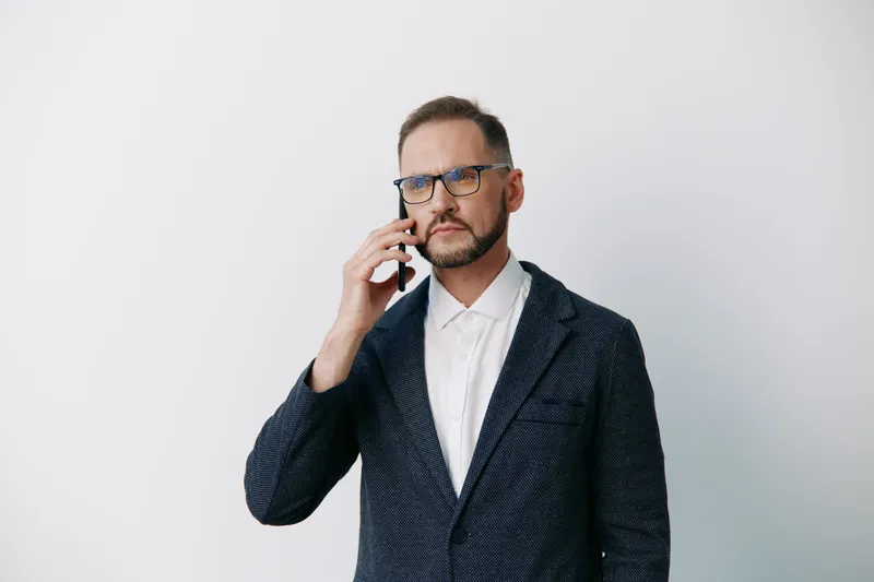A professional man on a call stands against a clean colored background, exuding focus, confidence, and calm while delivering concise business communication.