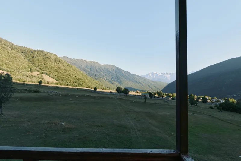 A serene landscape is seen through a window, revealing rolling valley plains and distant mountains under a clear sky. The window frame adds depth, inviting quiet contemplation of nature’s calm beauty.