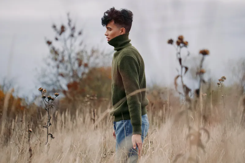 A solitary man stands in a tall grass field wearing a green knit turtleneck, conveying authenticity and credibility through natural light and quiet presence.