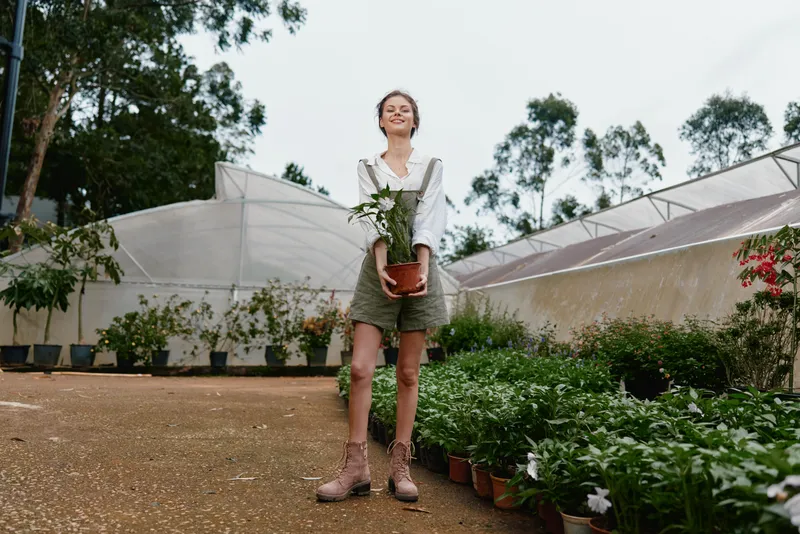 Young woman in a greenhouse holding a potted plant, smiling joyfully, dressed in a casual outfit, nature themed concept promoting gardening and plant care