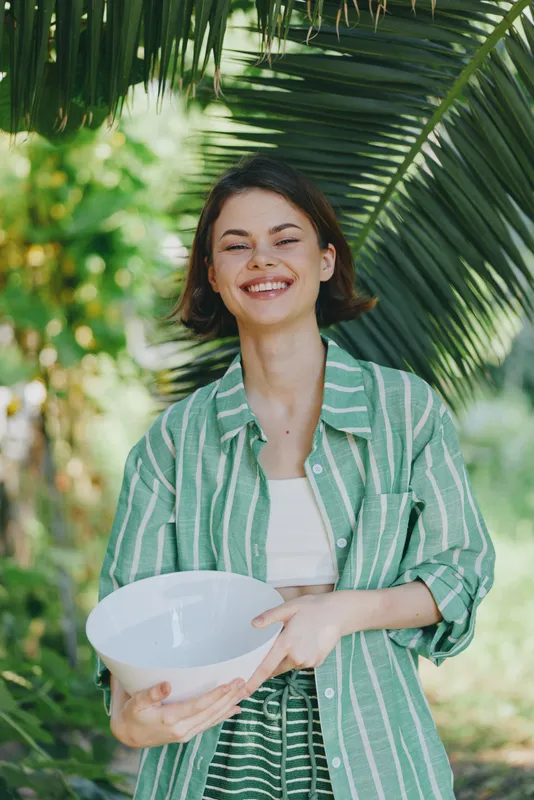Bright portrait of a cheerful woman outdoors, holding a white bowl, wearing a green striped jacket and white top, with lush tropical leaves in the background.