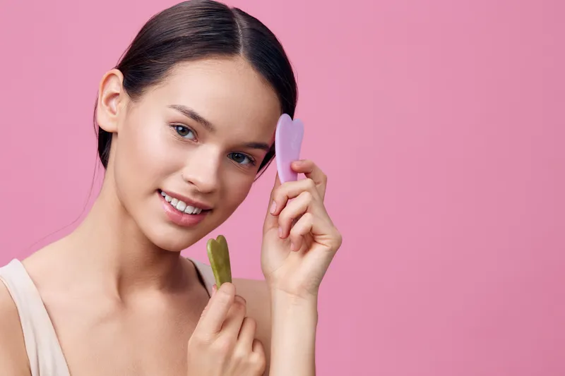 Young woman holding green and purple beauty tools, smiling with a soft expression against a pink background, illustrating skincare and self care concepts
