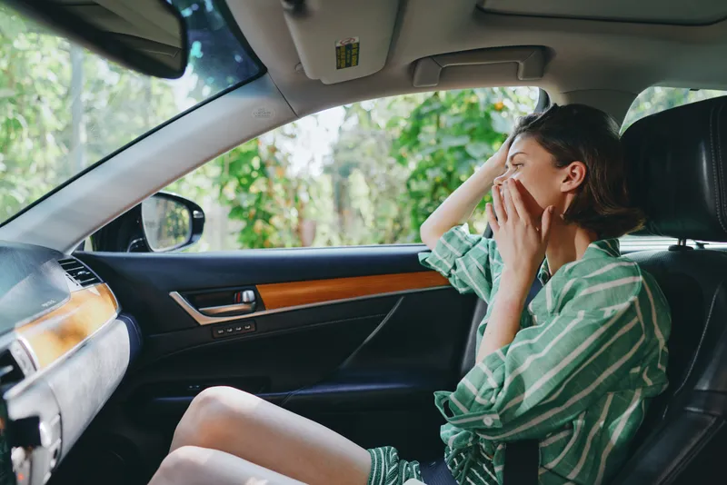A woman sits in a car, hands to her face in stress or surprise, as sunlight streams through the window and the dashboard surrounds her.