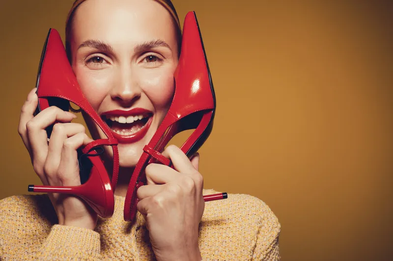 Smiling woman holds red high heel shoes near face, wearing yellow sweater. Studio portrait with warm background. Concept of fashion, style, and joyful emotions.