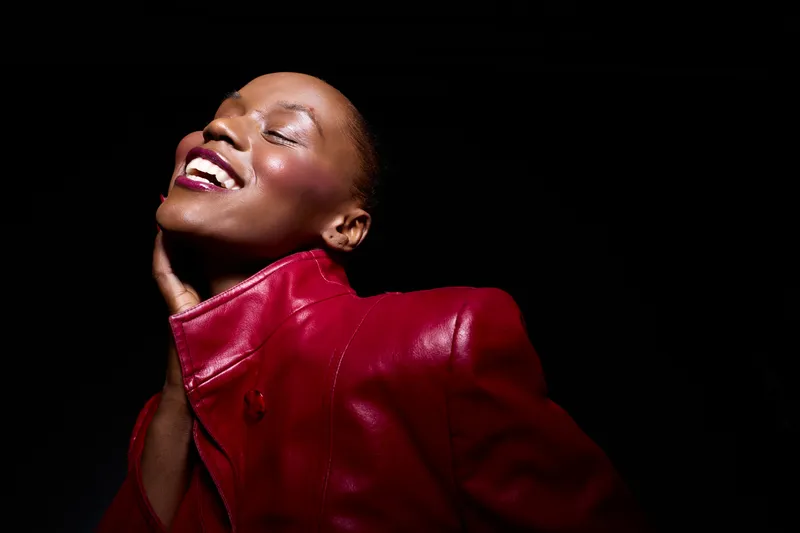 Portrait of a happy dark skinned African woman wearing a red leather jacket smiling with closed eyes, isolated on black background. Concept of joy, confidence, beauty, and fashion.