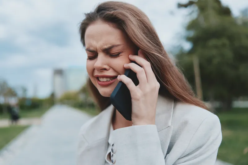 Young woman in beige jacket upset talking on phone outdoors, emotional distress, natural background, spring season, psychological problems concept