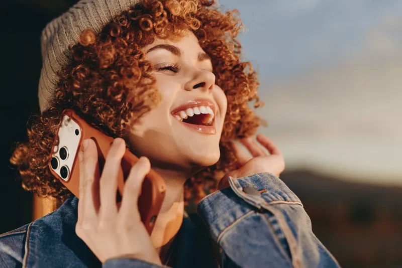 Woman lifestyle portrait with curly hair and denim jacket smiling while holding smartphone to her ear outdoors for a joyful call and communication in warm sunlight soft glow