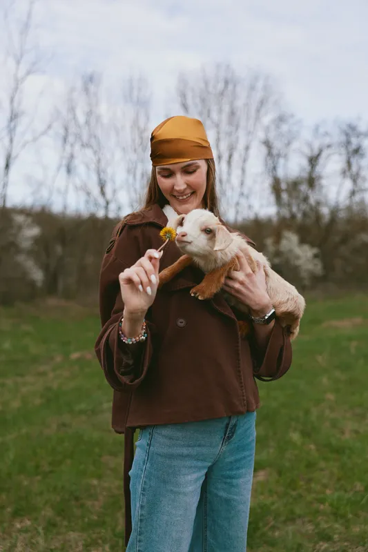 Woman in brown sweater and mustard headscarf holds a flower toward a curious goat kid in a sunny meadow, sharing a warm outdoor moment with nature and joy