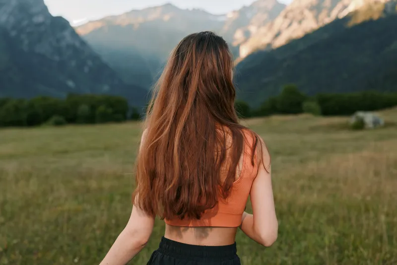A woman stands in a sunlit meadow, facing distant mountains. Her long hair falls over her back as she gazes across the open landscape, evoking calm, serenity, and a sense of freedom in nature.