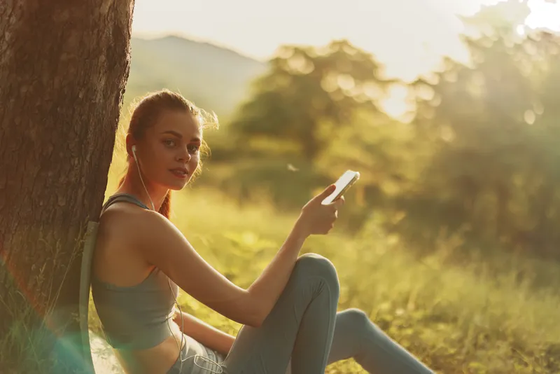 Young woman using smartphone outdoors in a natural setting, wearing casual sportswear, enjoying the moment, relaxed atmosphere, golden hour light