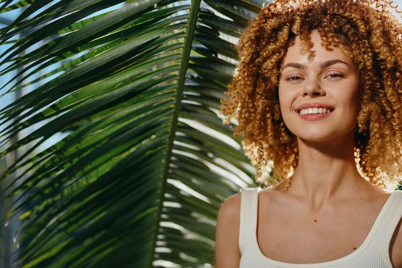 smiling woman with curly hair stands near tropical palm leaves on sunny day, wearing white top, natural light highlights her cheerful expression and vibrant hairstyle.