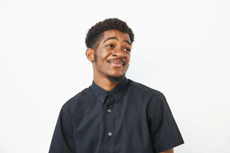 A cheerful African American man with a playful smile, wearing a black shirt, stands against a white background, radiating positivity and charm in this lively image.