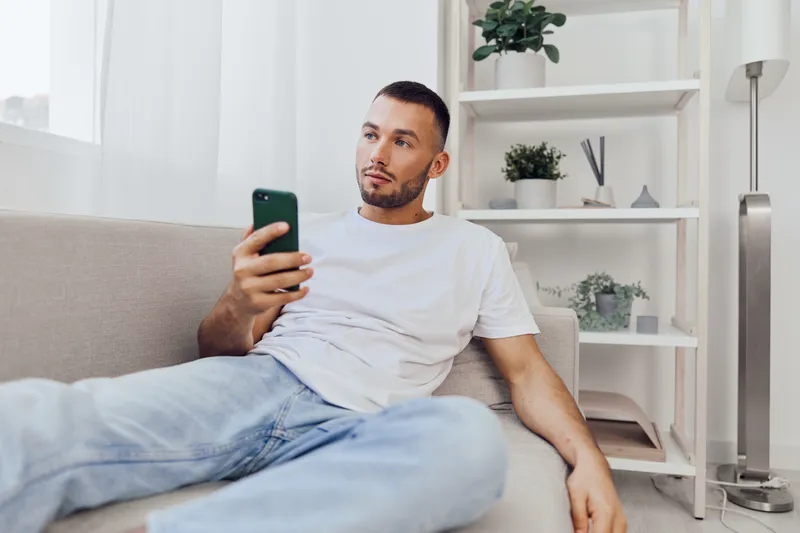 Thoughtful man sitting on a couch, casually dressed in a white t-shirt and jeans, engaged with his smartphone in a bright, modern interior.