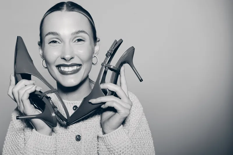 Smiling white woman with pale skin holds red high heels near face. Studio portrait with joyful mood and fashion concept on plain gray background.