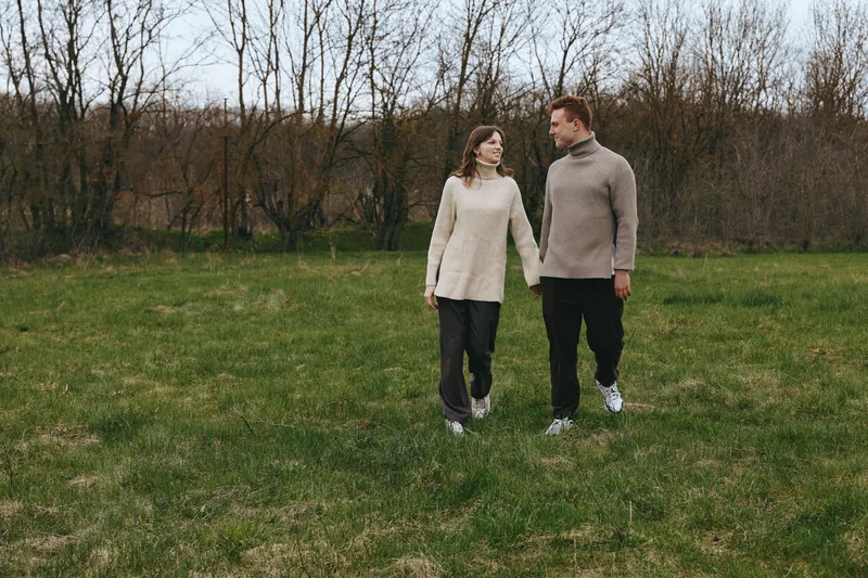 Couple walks hand in hand through a grassy park, wearing cozy sweaters, enjoying a calm outdoor moment, connected and content, with bare trees and soft light in the background, illustrating intimate