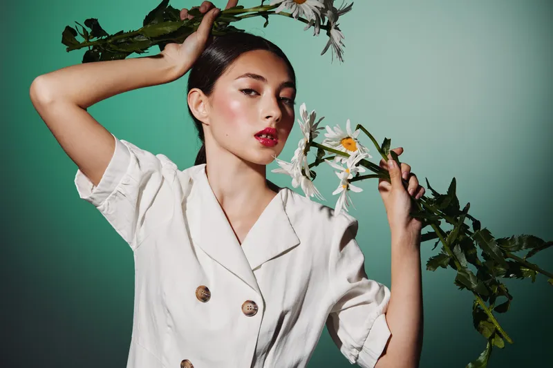 Young woman with dewy skin wears stylish white jacket holding floral branch near her face on turquoise background. Concept for beauty, creative fashion, skincare, elegance, and floral accents.