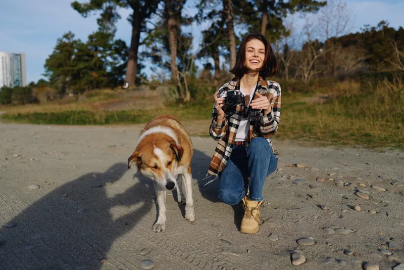 Woman kneeling on a pebble beach with her dog, smiling and holding a camera, casual outfit, clear sky, grassy backdrop, outdoor photography moment on a sunny day