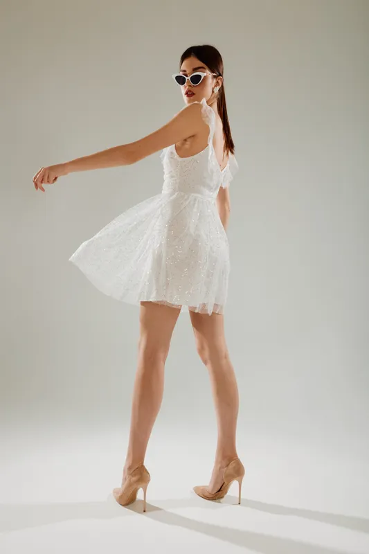Young woman in a white lace dress and sunglasses poses confidently on a light background. Concept of beauty, fashion, elegance, and summer style in studio lighting.