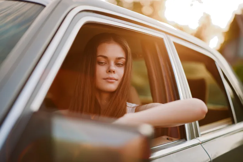 Young woman enjoying a summer drive in a stylish car, showcasing relaxation and joy against a vibrant sunset backdrop.