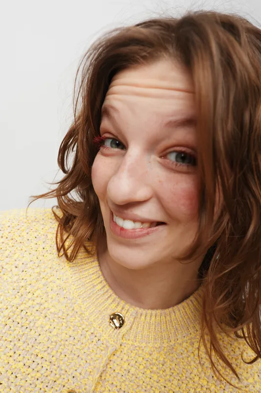 Smiling woman with curly brown hair wearing yellow sweater looks at camera with playful expression on white background. Concept of happiness, casual style, and natural beauty.