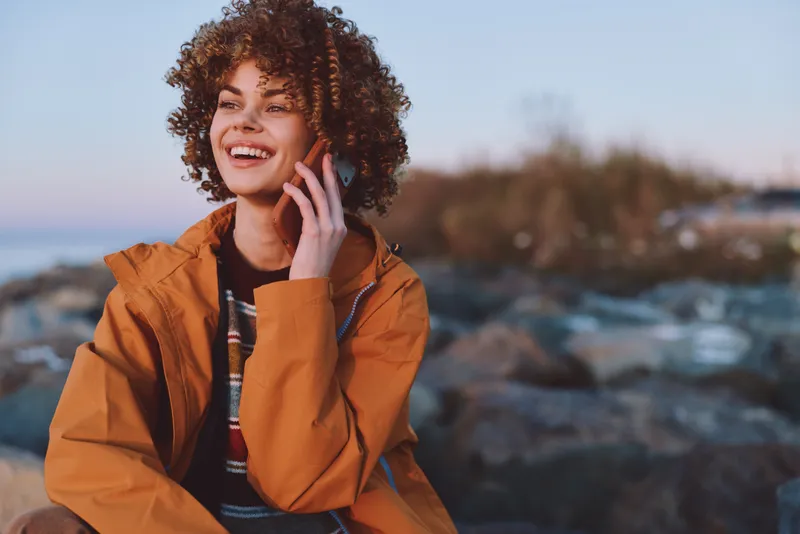 Smiling woman lifestyle portrait, curly hair and orange jacket outdoors, talking on phone for communication by the coast, calm ocean and soft sunset light