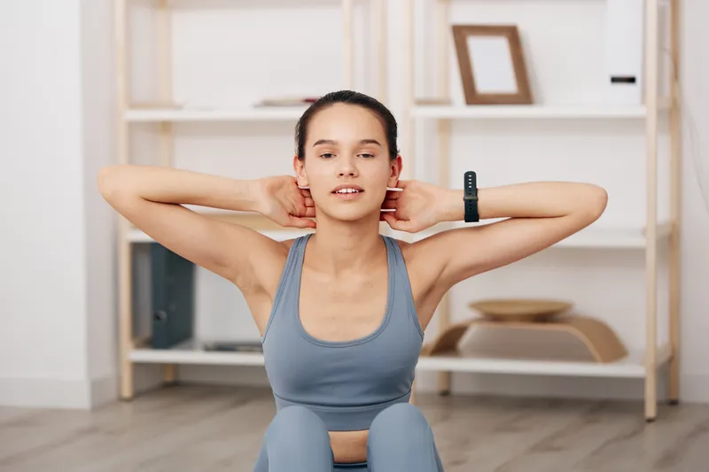 Woman in athletic wear performs a neck stretch during a home workout, showcasing fitness, balance and focus in a tidy room with shelves and simple decor.