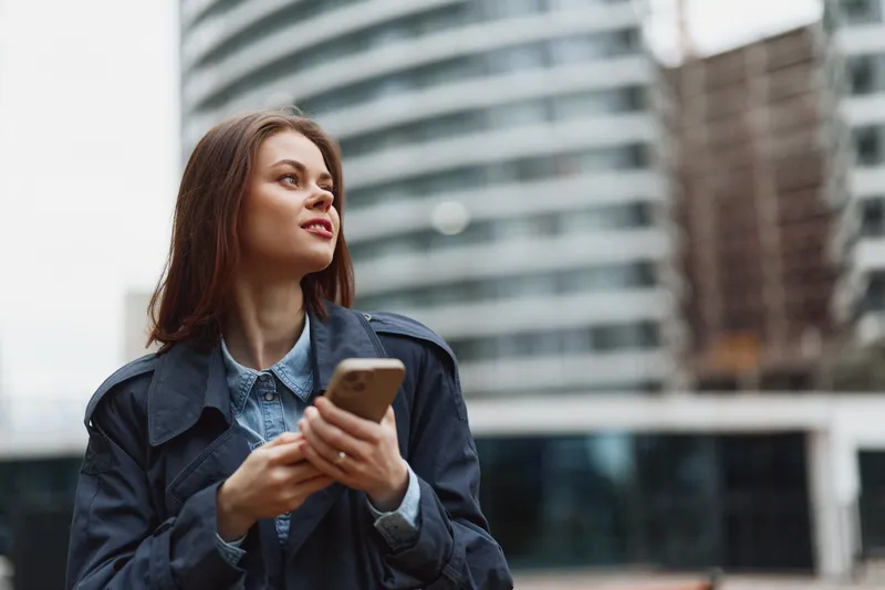 A young female urban professional stands outdoors, holding a smartphone and looking up, wearing a denim jacket and collared shirt, conveying contemporary city life and curiosity.