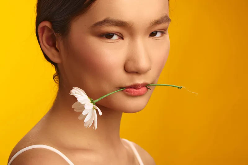 An Asian woman holds a delicate flower between her lips while standing before a vibrant yellow backdrop, conveying a serene, confident, and stylish presence.