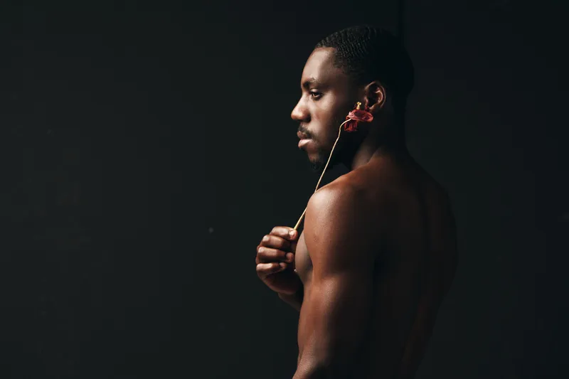 Creative conceptual portrait of African man with bare torso holding flower near face against dark background in visual artistic style. Black skin, profile and minimalistic composition.