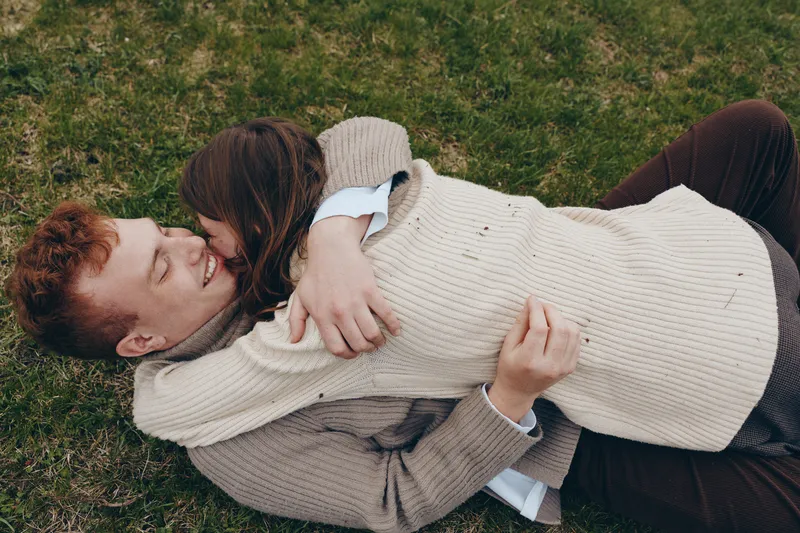 A couple shares a warm embrace on a grassy lawn, relaxed and smiling in cozy knitwear. A candid lifestyle moment highlighting affection, comfort, and intimate connection outdoors.