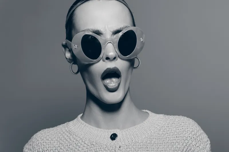 Surprised white woman with pale skin wearing large round sunglasses and hoop earrings looks shocked isolated on grey background. Concept of fashion and emotional expression.