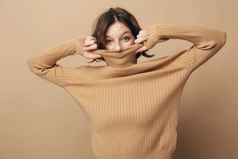 Young woman in beige knitted sweater pulls collar over face, peeking with playful eyes. Studio portrait on neutral background. Concept of cozy fashion and casual style.