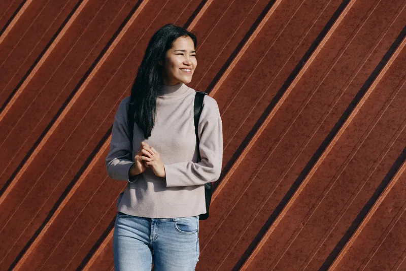 Casual woman with backpack stands outdoors beside a diagonal wooden wall, smiling, showcasing everyday style, relaxed pose, and urban lifestyle photography for fashion and travel scenes