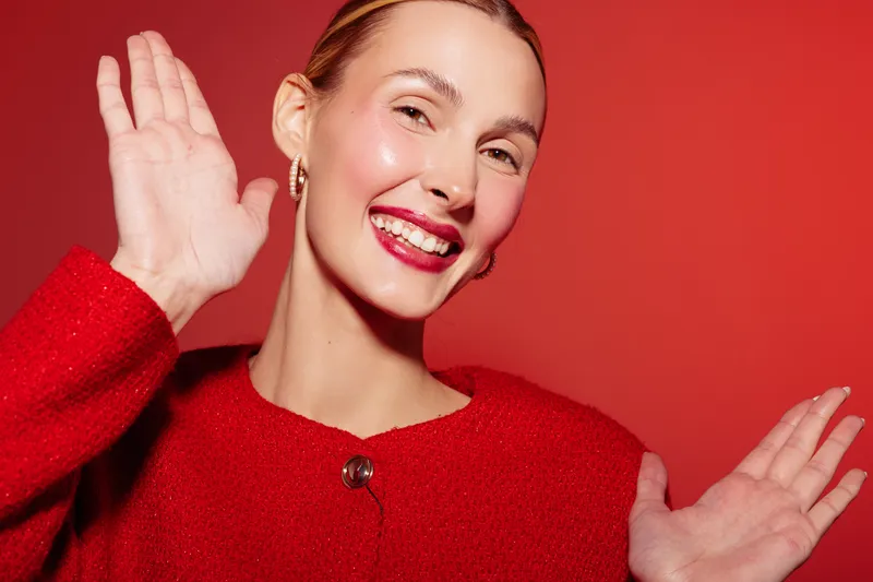 Smiling woman in red sweater waves hands with joyful expression on red background. Close-up portrait with bright makeup and earrings. Concept of happiness, celebration, and positive emotions.