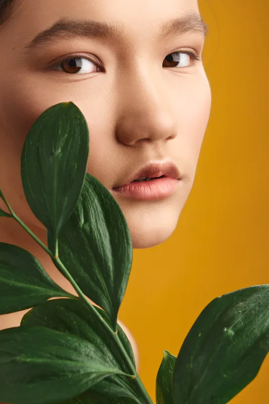 asian woman with calm expression peers beside lush green leaves against a warm background, conveying natural grace, simplicity, and contemplative mood in a close up portrait.