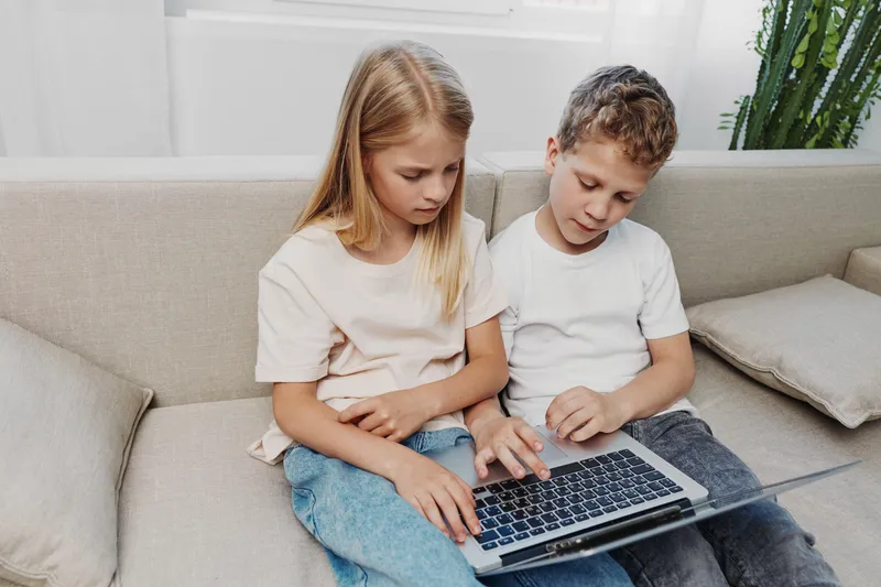 Two focused children sitting closely on a beige couch using a laptop together in a bright living room. Concept of learning, technology, cooperation, and childhood curiosity.