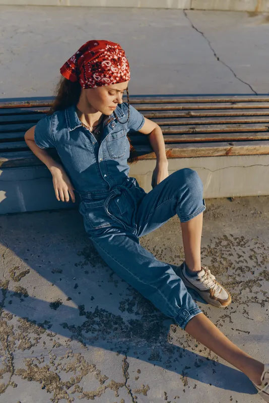 lifestyle woman in modern boho-western denim street style sits relaxed on a concrete surface wearing a red bandana in warm film color tones