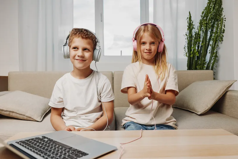Two children wearing headphones sit on a couch at home, enjoying music or audio content. A laptop and indoor plants are visible, creating a cozy family atmosphere.